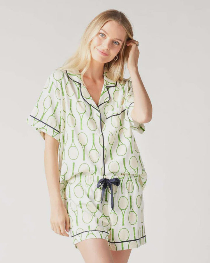 A woman smiles at the camera, wearing the Zertz TENNIS TIME PAJAMA SHORTS SET—white with green tennis racket monograms—standing against a plain light background.