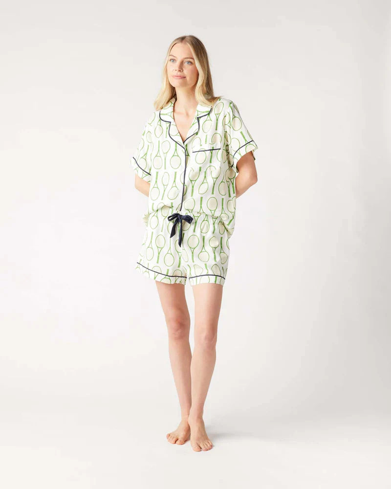 A woman stands barefoot against a plain background, wearing the Zertz TENNIS TIME PAJAMA SHORTS SET featuring a green tennis racket American Summer print with black trim. Her hands are in her shorts pockets and she has a relaxed expression.