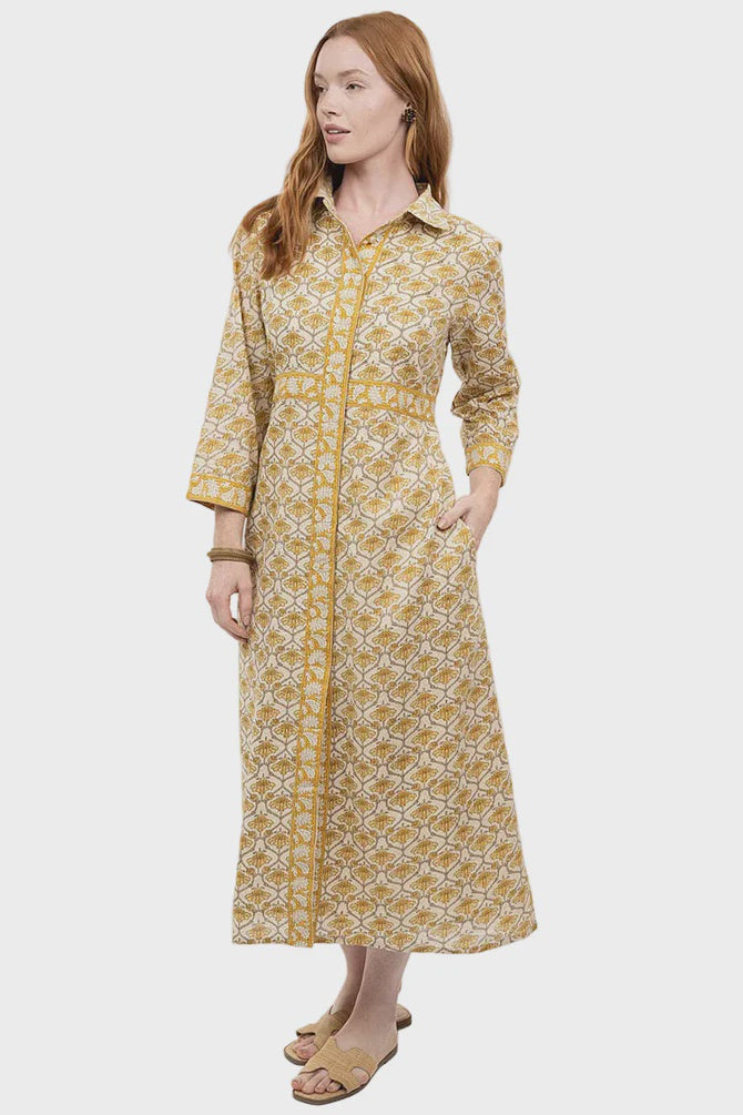 A woman models the Zertz DOUBLE BUTTON SHIRT DRESS, a long-sleeved, ankle-length yellow and white piece with floral details. She stands with one hand in her pocket, the other by her side, finishing the look with tan sandals.