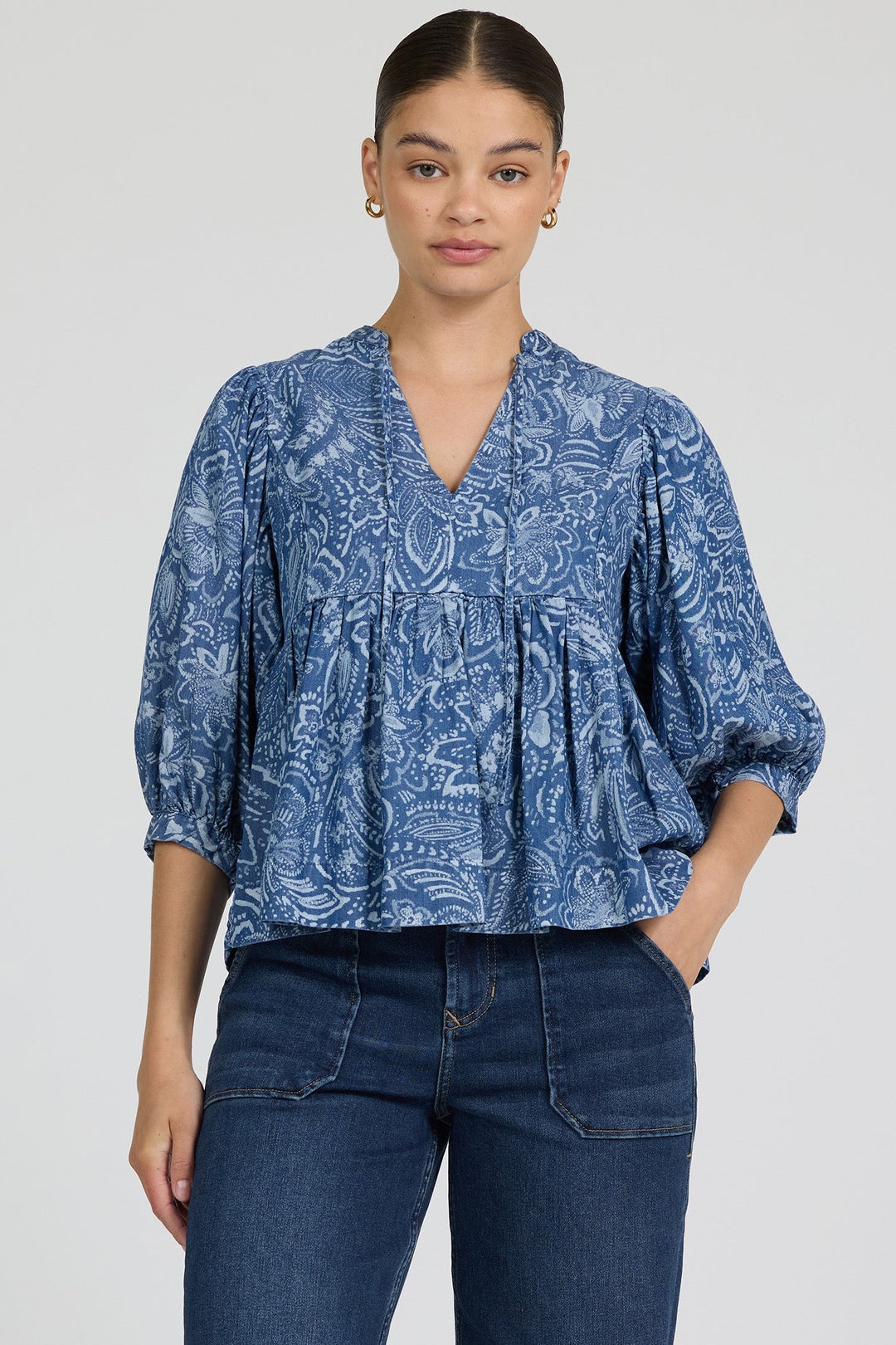 A woman wearing the Zertz MALIA TOP—a feminine, floral blue blouse with three-quarter sleeves made from breathable fabric—and dark blue jeans stands against a plain light background, looking at the camera with a neutral expression.