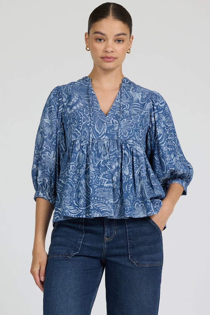 A woman wearing the Zertz MALIA TOP—a feminine, floral blue blouse with three-quarter sleeves made from breathable fabric—and dark blue jeans stands against a plain light background, looking at the camera with a neutral expression.