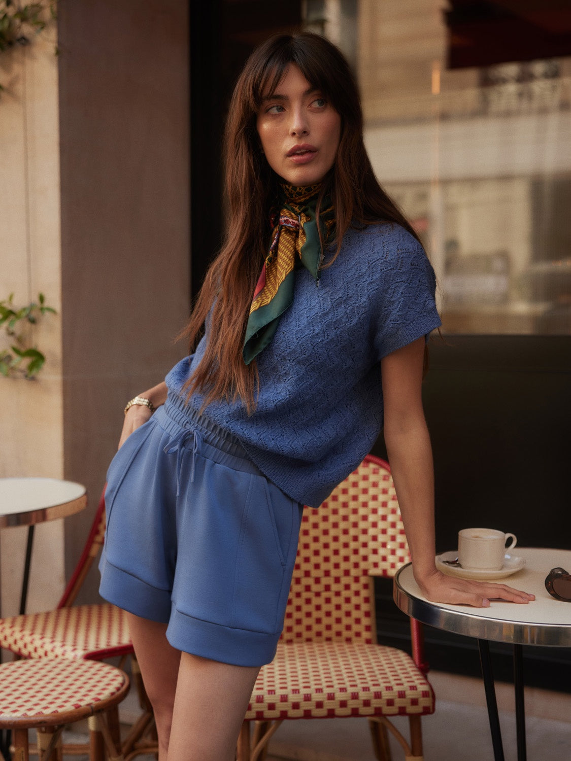A woman with long brown hair wears the Zertz ATRIUM HIGH RISE SHORT 4.5 INSEAS, a textured classic blue top, and a colorful scarf as she stands by an outdoor café table—creating a chic summer look among red and white chairs.