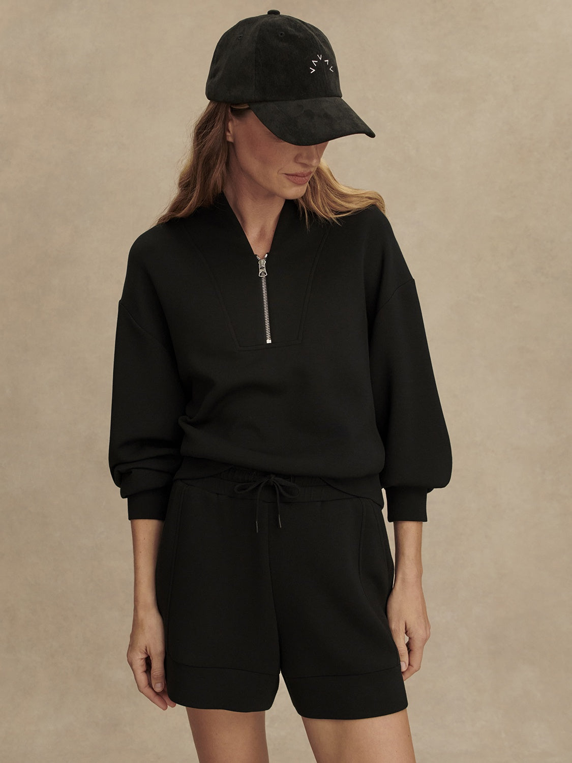 A woman wears the Zertz ATRIUM HIGH RISE SHORT 4.5 INSEAM BLACK with a black zip-up sweatshirt and baseball cap, standing against a beige background and looking down to her left, hands relaxed by her sides.