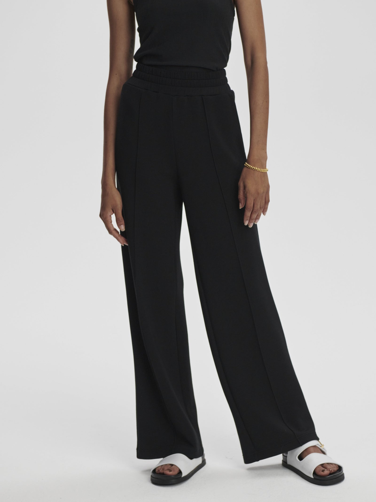 A person wearing a black sleeveless top, Zertz THE WIDE LEG PANT BLACK, a gold bracelet, and black-and-white sandals stands against a plain white background.