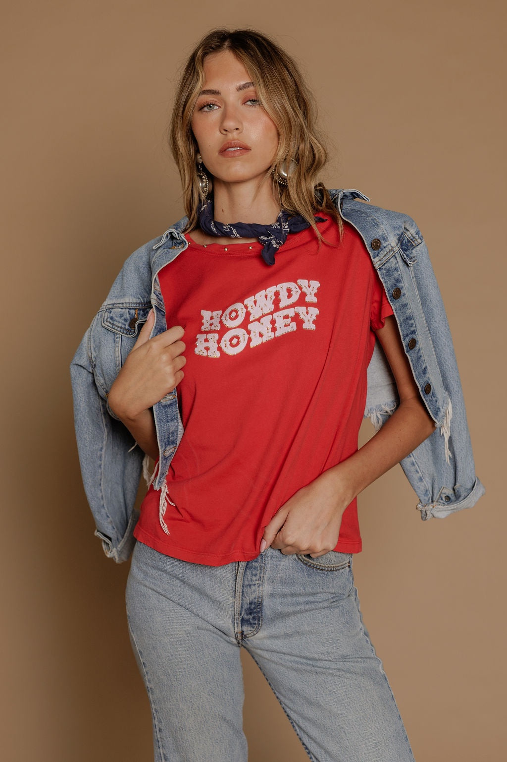 A woman in a red Zertz HOWDY HONEY BOY TEE, light blue jeans, and a blue bandana poses against a beige background, accessorized with delicate necklaces and a denim jacket draped over her shoulders.