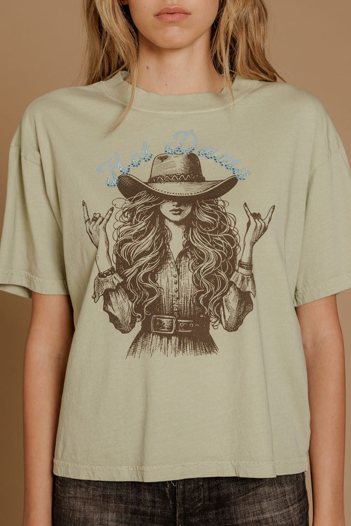 A woman wears the Zertz HOT DAMN WIDE BOY TEE, a sage green relaxed fit T-shirt featuring an illustrated cowgirl with long wavy hair, a wide-brim hat, and hand signs. Her face is partly out of frame.