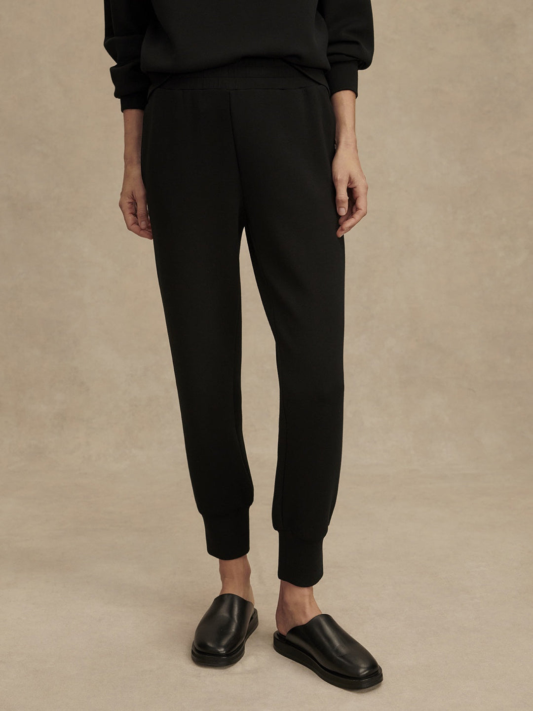 Against a beige background, a person wears Zertz THE SLIM CUFF PANT 25-BLACK with tapered legs, a matching black top, and slip-on shoes. Cropped from the shoulders down, these versatile joggers are a true wardrobe staple.