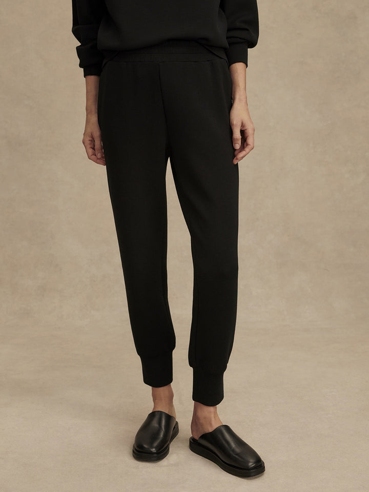 Against a beige background, a person wears Zertz THE SLIM CUFF PANT 25-BLACK with tapered legs, a matching black top, and slip-on shoes. Cropped from the shoulders down, these versatile joggers are a true wardrobe staple.