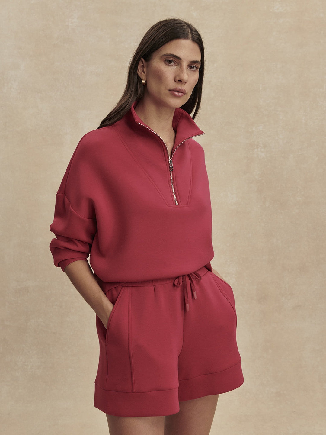 A woman with long brown hair models the Zertz ATRIUM HIGH RISE SHORT 4.5 INSEAM in Rich Raspberry, paired with a matching pullover, standing with hands in pockets against a beige background.