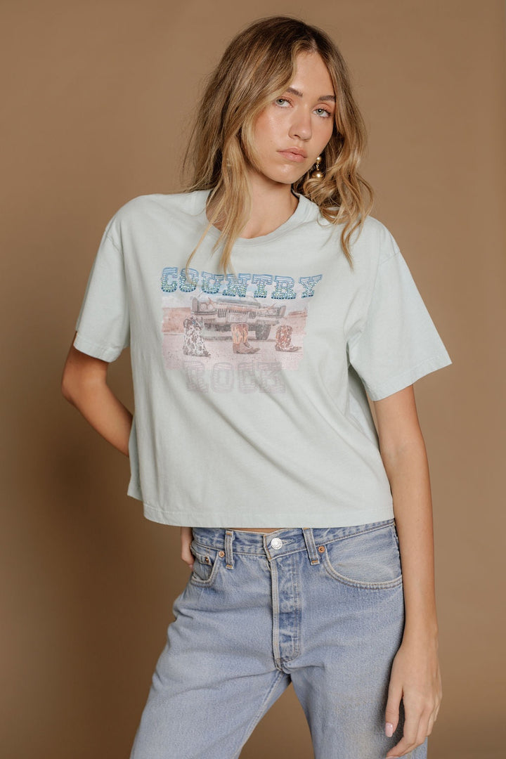 A young woman wears the Zertz COUNTRY ROCK WIDE BOY TEE, a premium cotton blend in light blue with a vintage truck and COUNTRY graphic, paired with high-waisted light-wash jeans against a tan background.