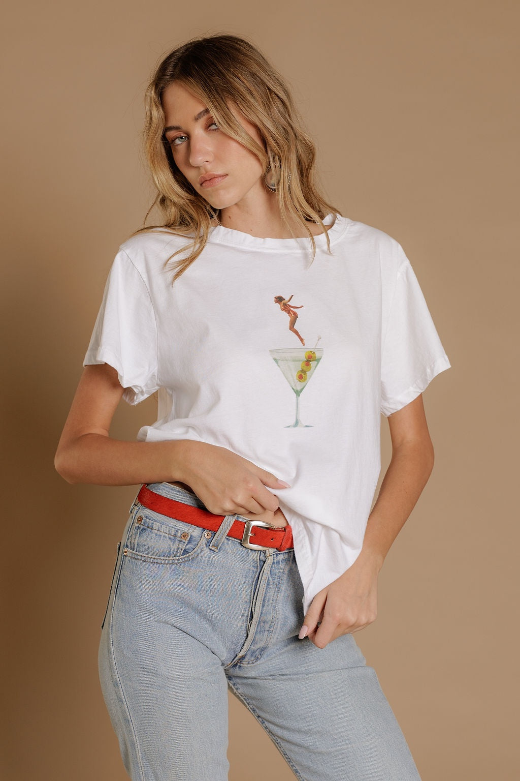 A woman with wavy blonde hair stands against a beige background, wearing Zertz's DIP INTO MARTINI TEE with light blue jeans and a red belt—an ideal choice for cocktail lovers adding fun style to any casual look.