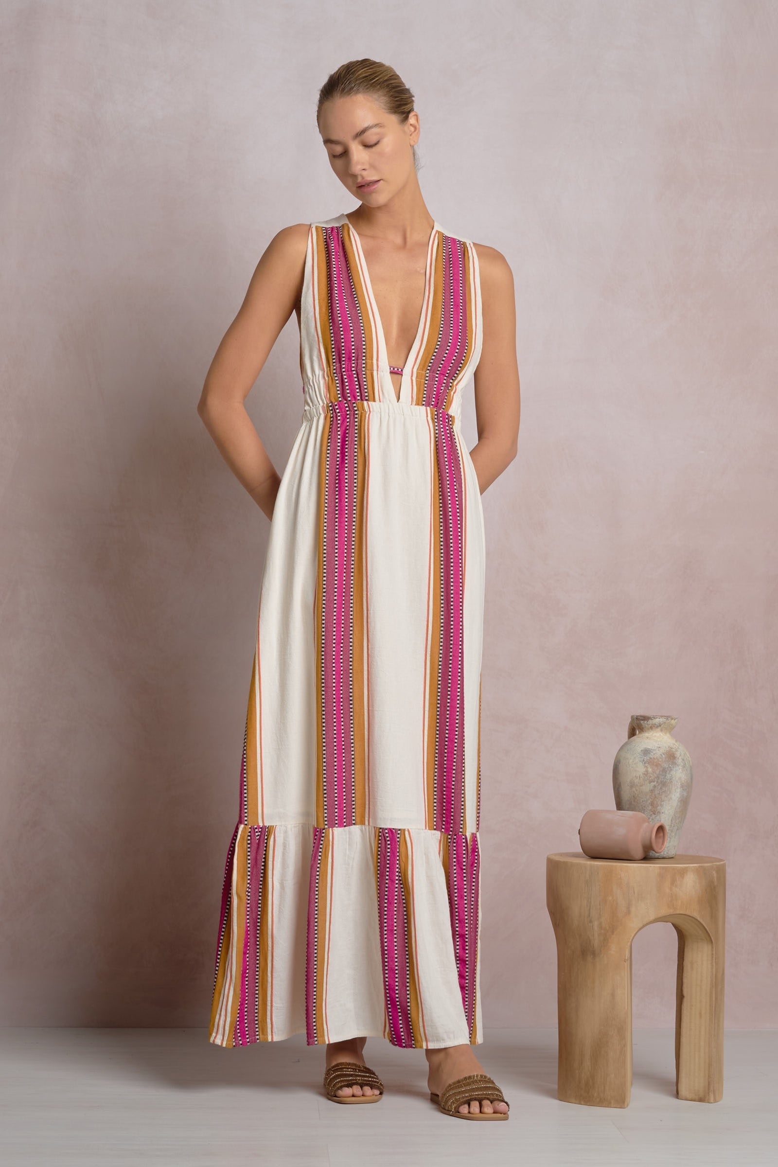 A woman wears the Zertz RUFFLE MAXI HALTER DRESS, a sleeveless white maxi with pink, orange, and yellow vertical stripes and pockets. She pairs it with flat sandals. Beside her is a wooden stool holding two ceramic vases.