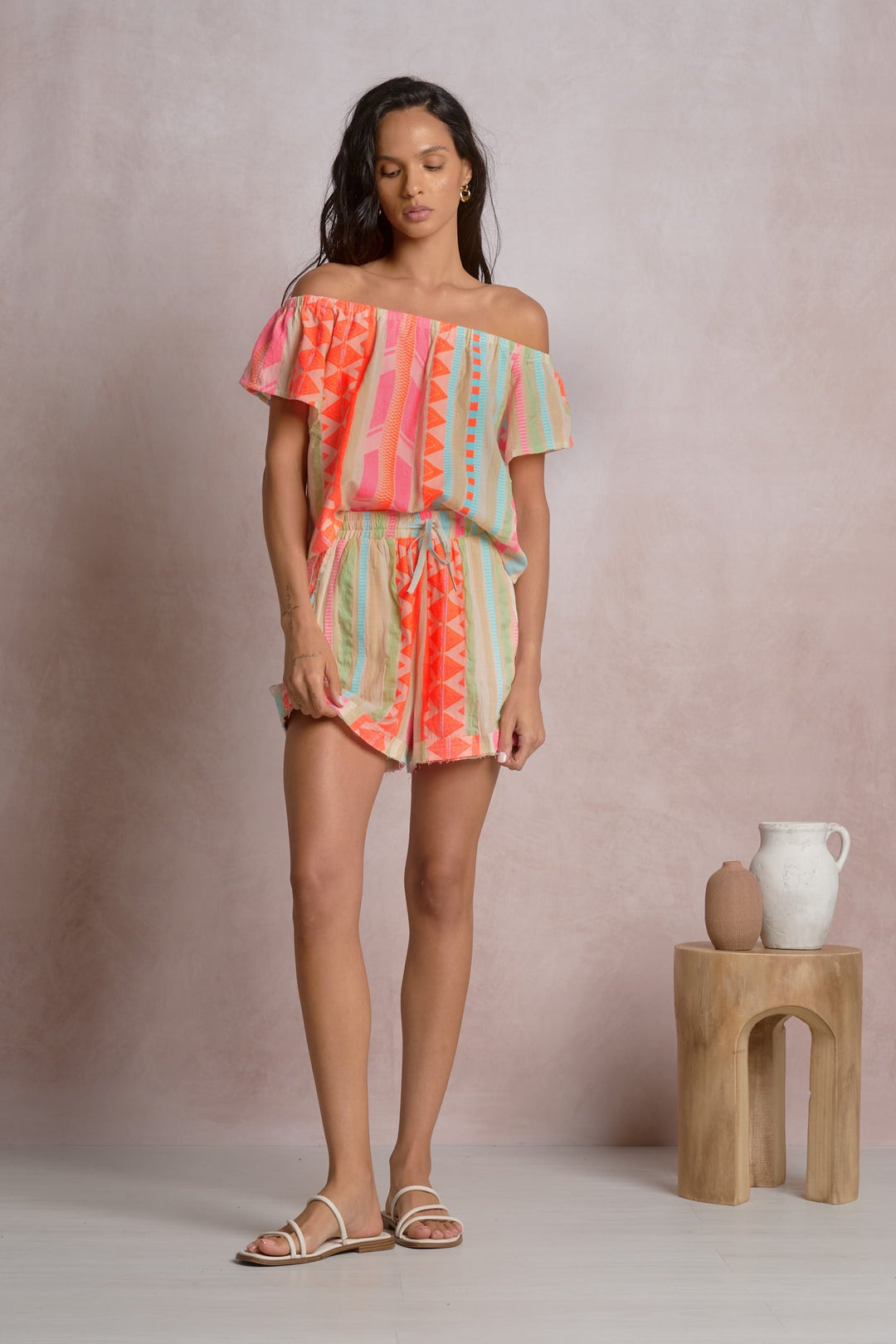 A woman poses indoors in the Zertz OFF SHOULDER RUFFLE SLEEVE TOP with geometric patterns, white sandals, and gold hoop earrings. She stands beside a small wooden stool holding two ceramic vases against a light pink backdrop.