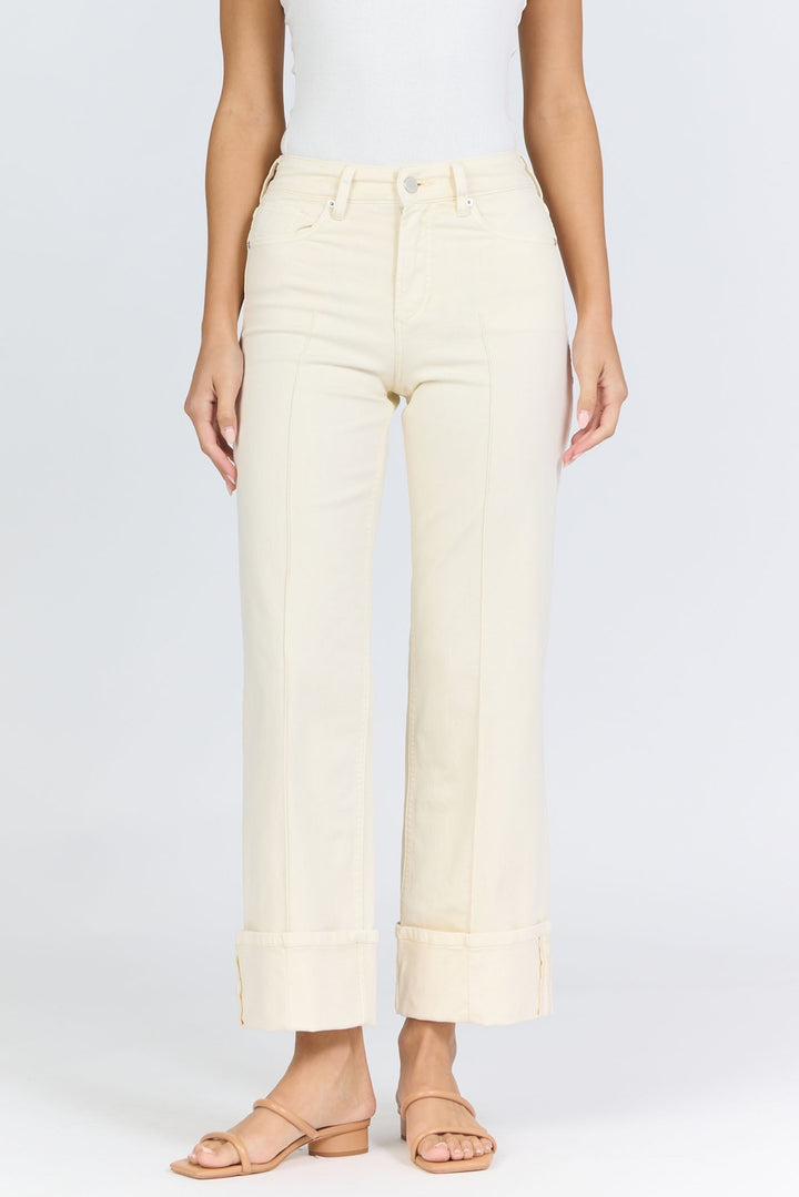 A person in Zertz’s HOLLY JEAN high-waisted, light beige wide-leg jeans with large cuffs, a white sleeveless top, and tan sandals stands against a plain white background in timeless style.