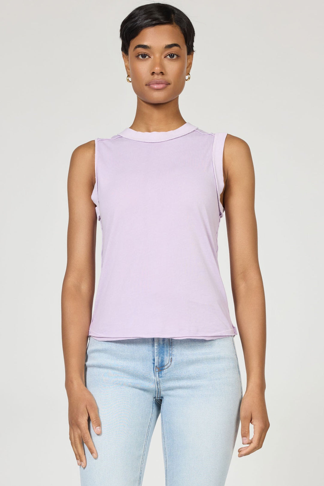 A woman with short dark hair wears the sleeveless light purple ZO TANK by Zertz and light blue jeans, standing against a plain background and looking at the camera—an essential wardrobe staple.