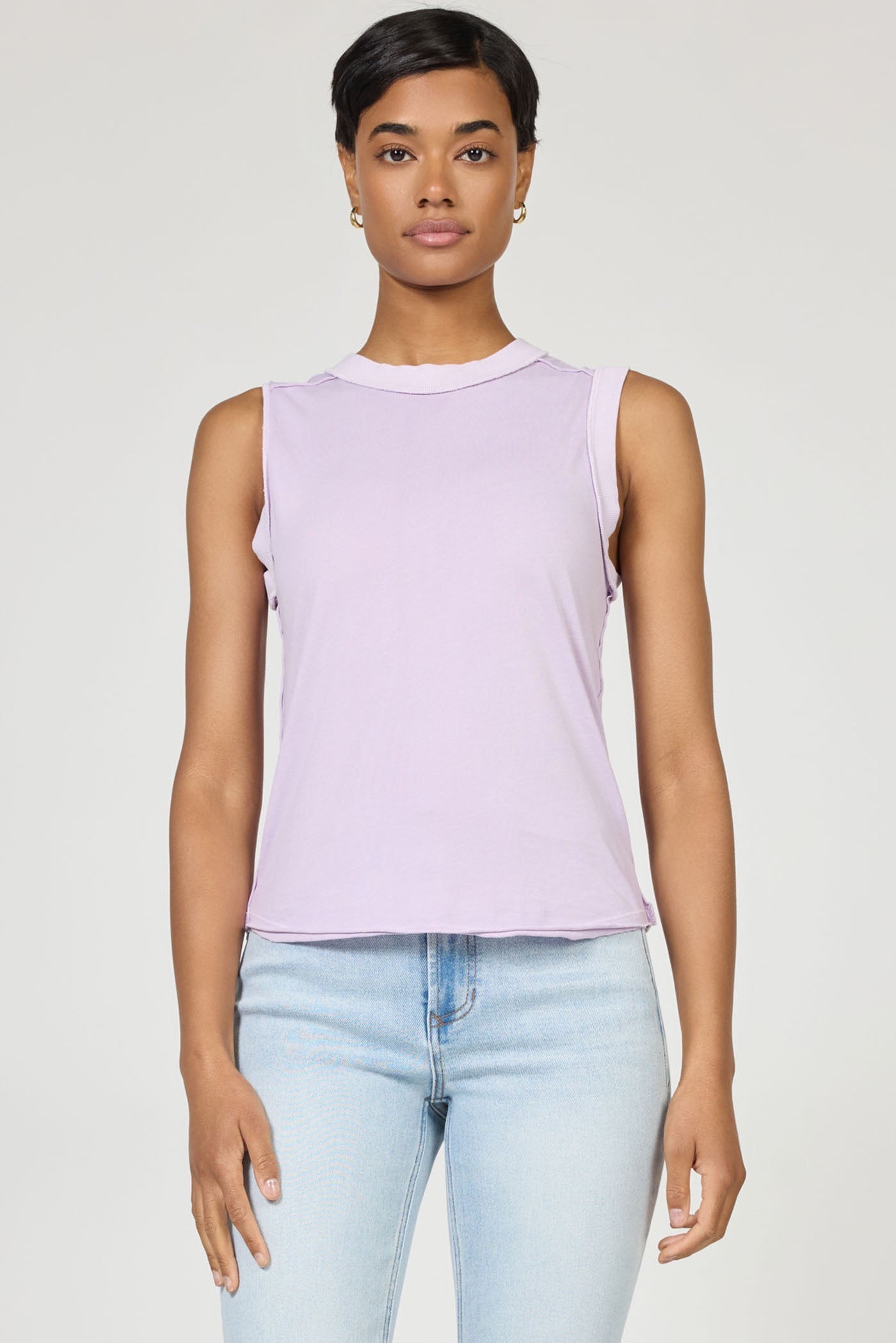 A woman with short dark hair wears the sleeveless light purple ZO TANK by Zertz and light blue jeans, standing against a plain background and looking at the camera—an essential wardrobe staple.