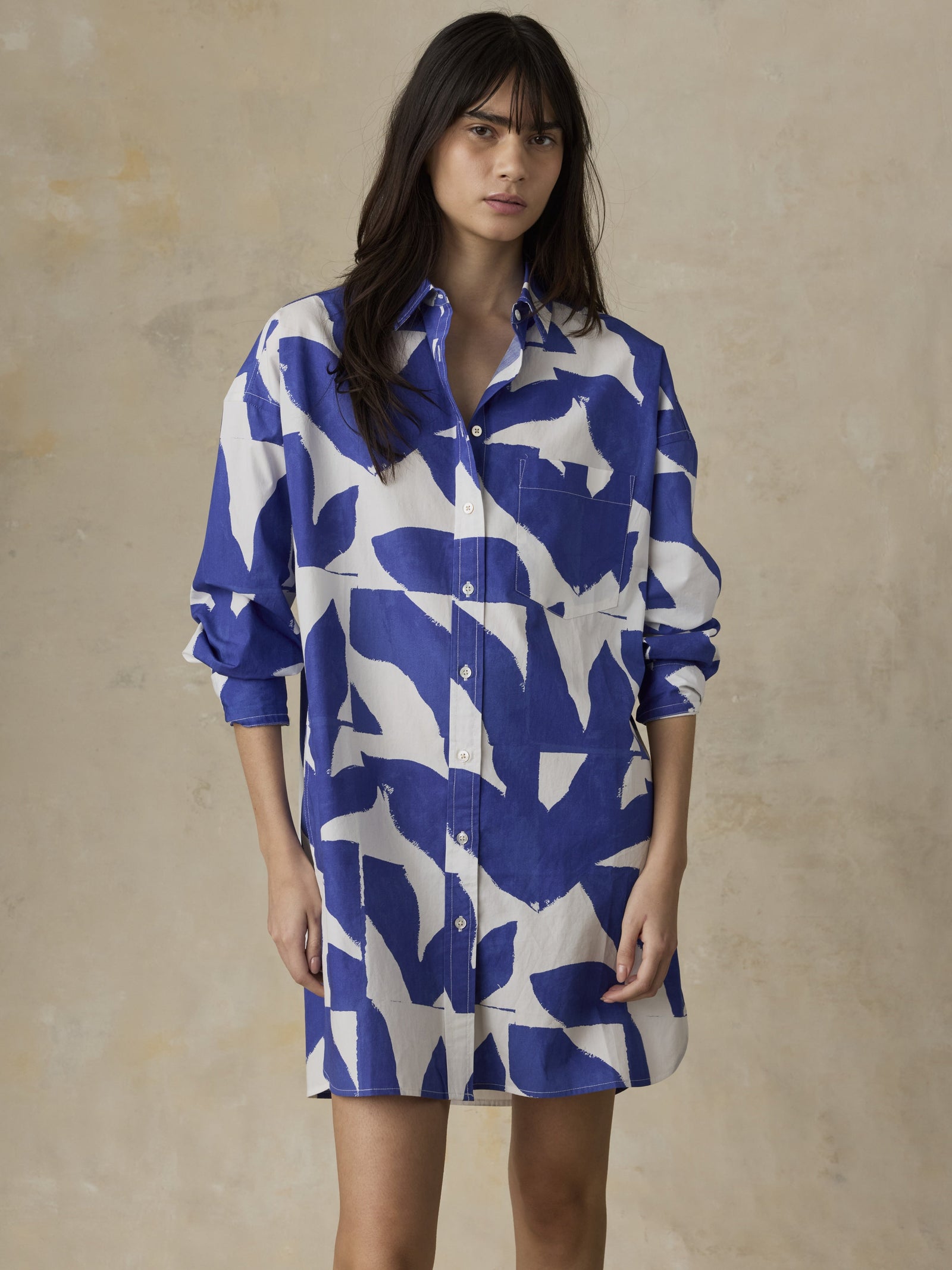Against a beige textured background, a woman in the Zertz COSTAL POPLIN KOVA SHIRTDRESS—featuring bold blue and white abstract patterns—stands with loose dark hair and relaxed arms, embodying effortless coastal elegance.