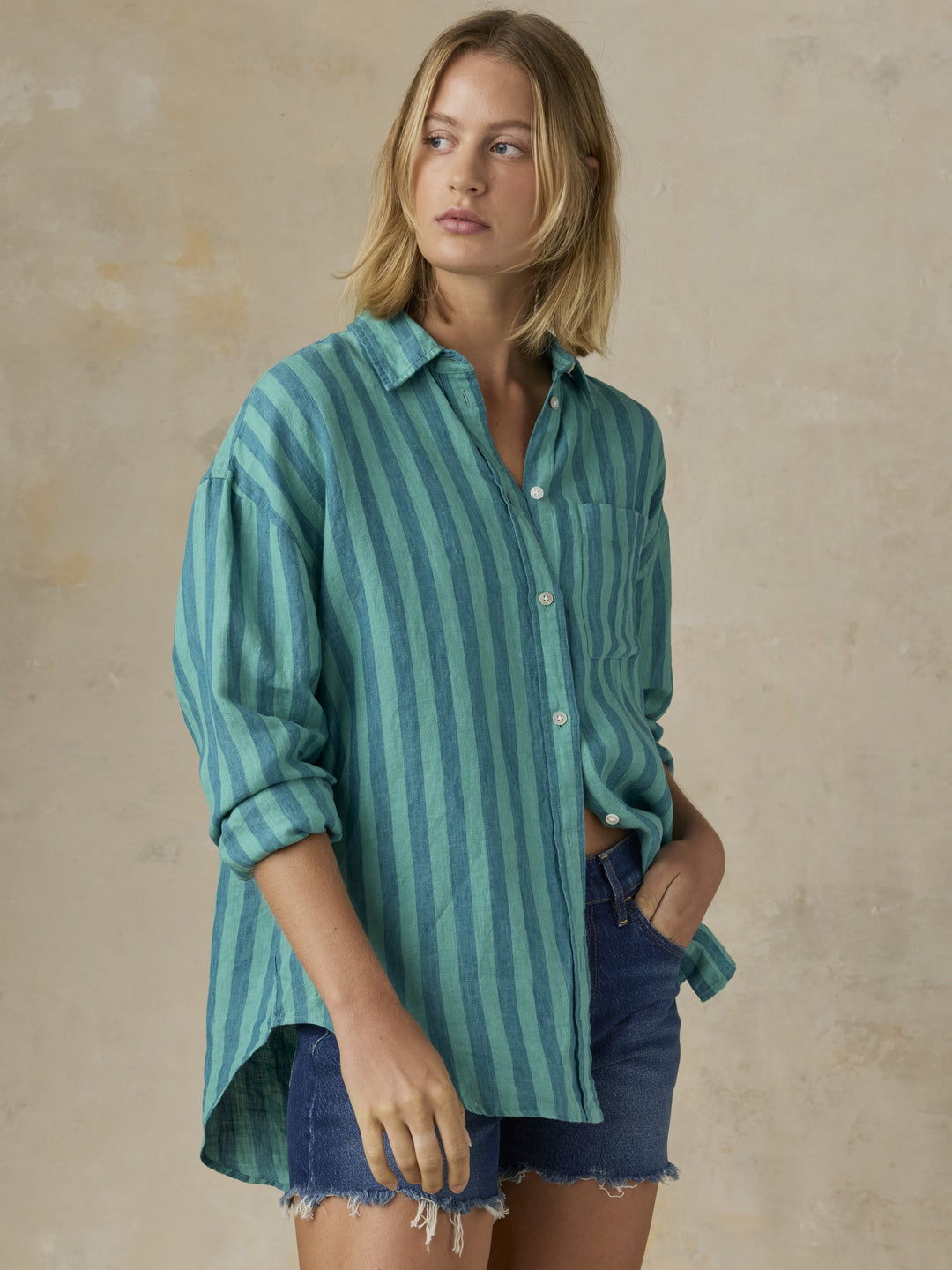 A woman with shoulder-length blonde hair models the Zertz LAGUNA LINEN RELAXED SHIRT in teal and blue stripes, sleeves rolled up, paired with dark denim shorts. One hand is in her pocket as she looks slightly to the side against a neutral background.