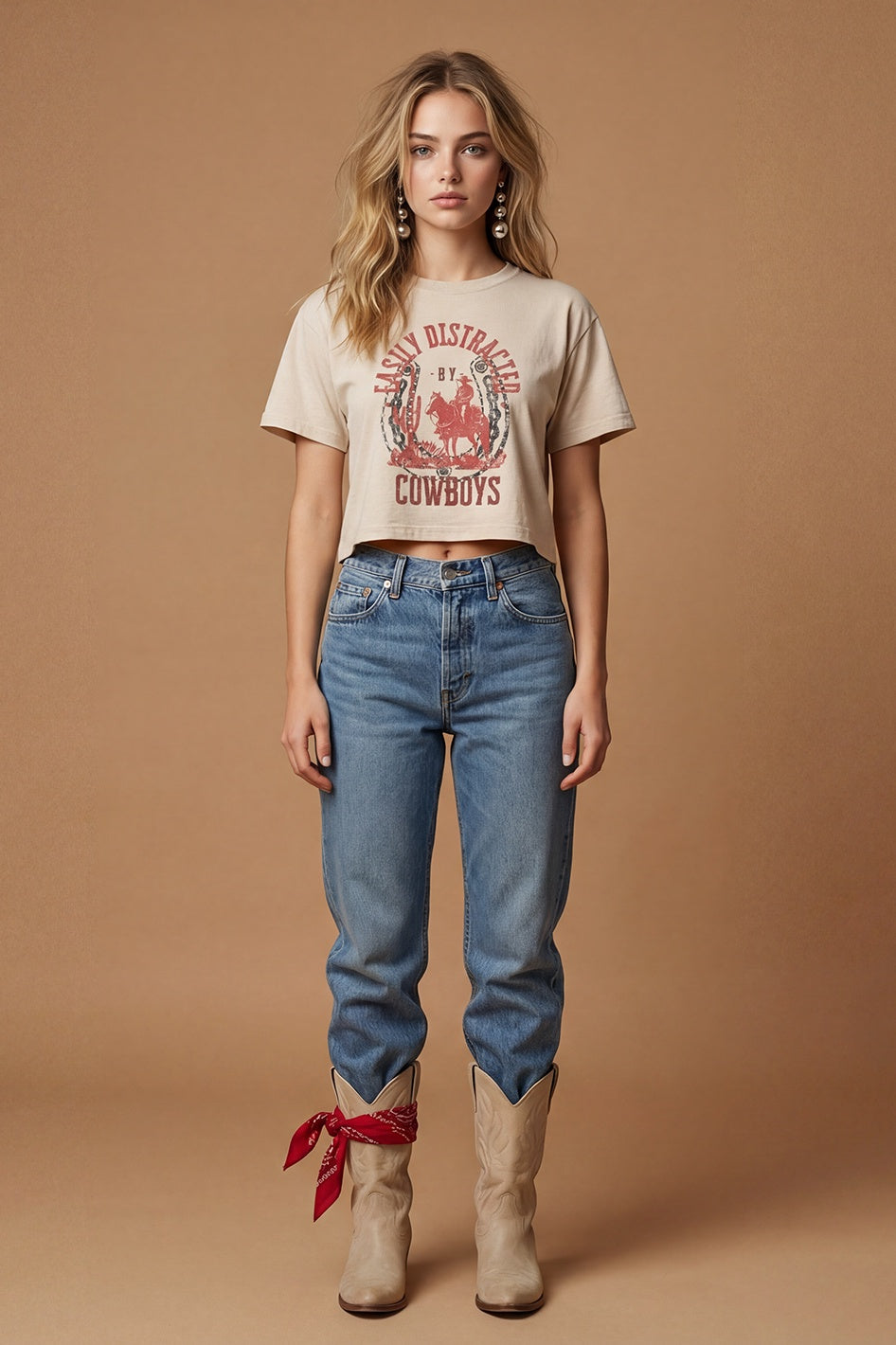 A young woman with long wavy blonde hair and a neutral expression stands against a tan backdrop, wearing the Zertz EASILY DISTRACTED WIDE BOY TEE with a cowboy graphic, blue jeans, tan cowboy boots, and a red bandana tied around her ankle.