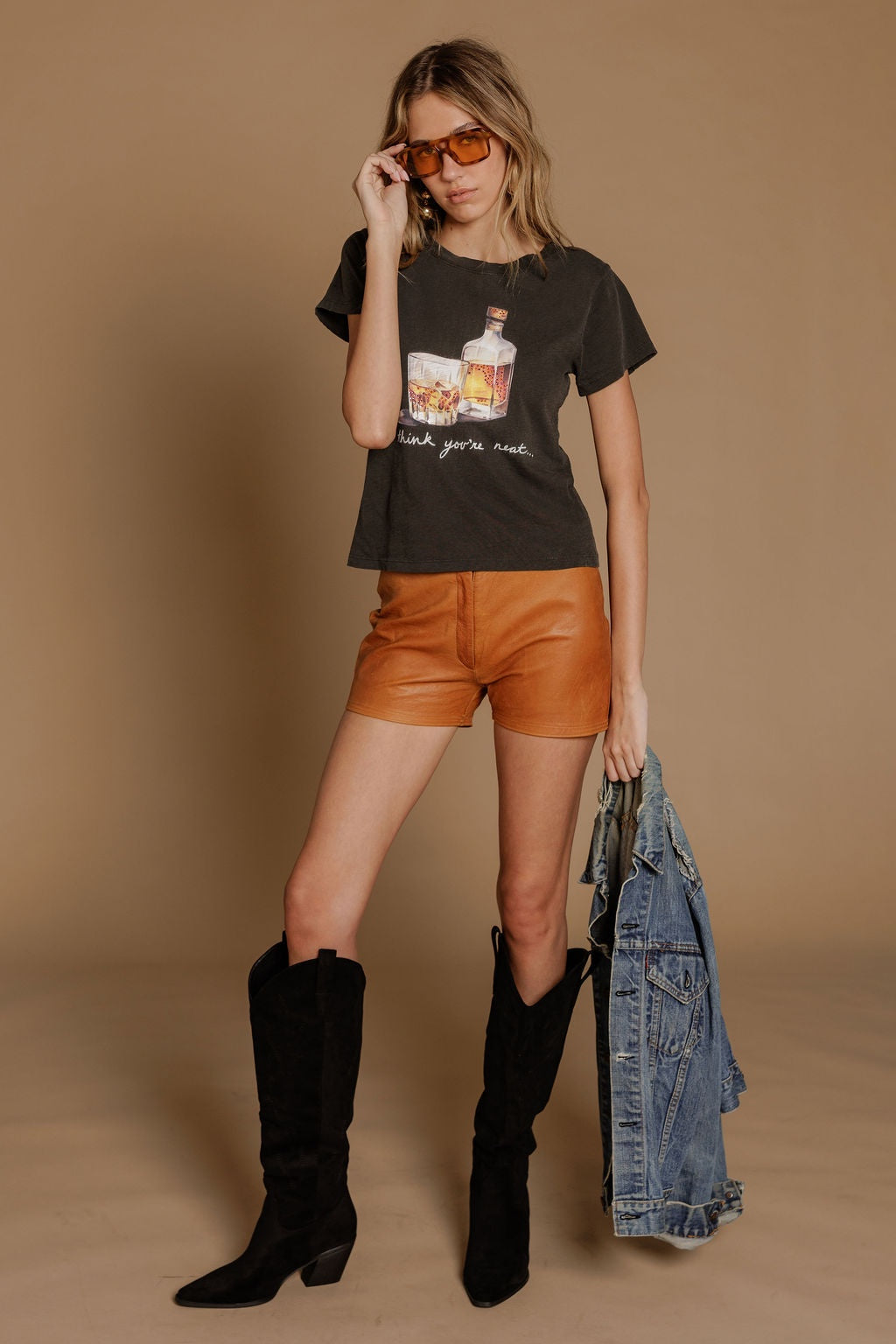 A woman poses against a tan background in orange sunglasses, tan leather shorts, black knee-high boots, holding a blue denim jacket, and wearing the Zertz I THINK YOUR NEAT SLIM TEE.