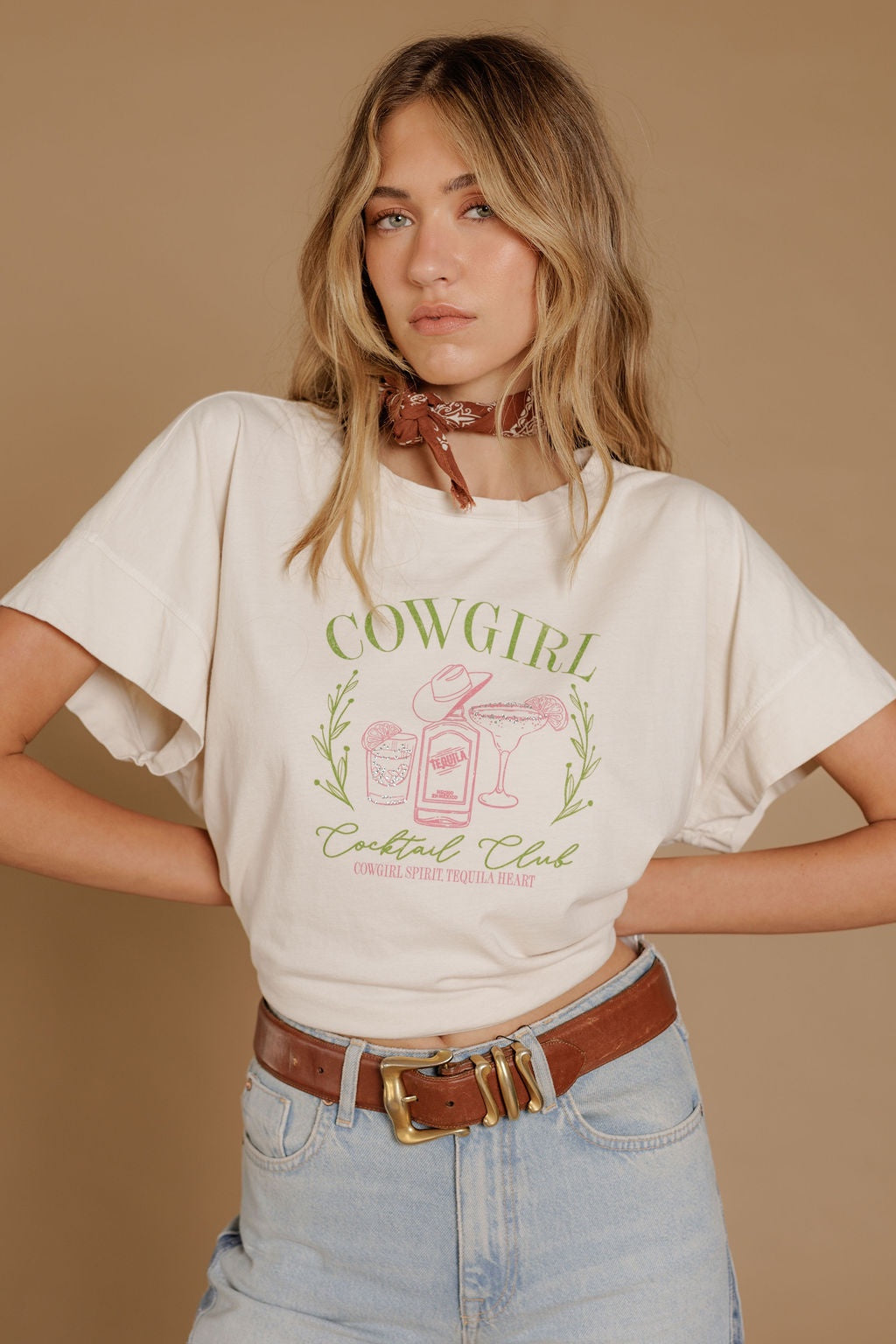 A woman stands against a beige background wearing the Zertz COWGIRL COCKTAIL HI LOW TEE in white, paired with light blue high-waisted jeans, a brown belt, and a brown patterned scarf tied around her neck.