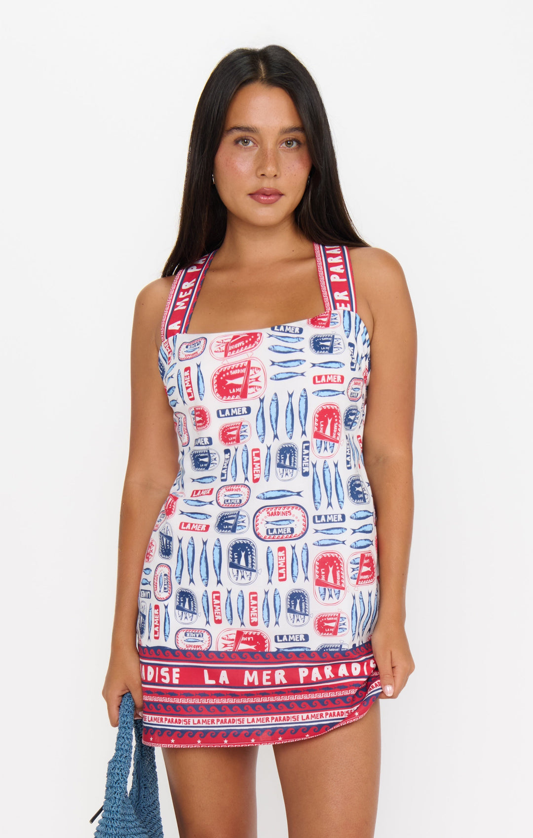 A woman with long dark hair wears the Zertz DETOUR MINI DRESS, a sleeveless dress featuring red, blue, and white graphic prints with thick straps. She holds a blue knitted bag and stands against a white background.