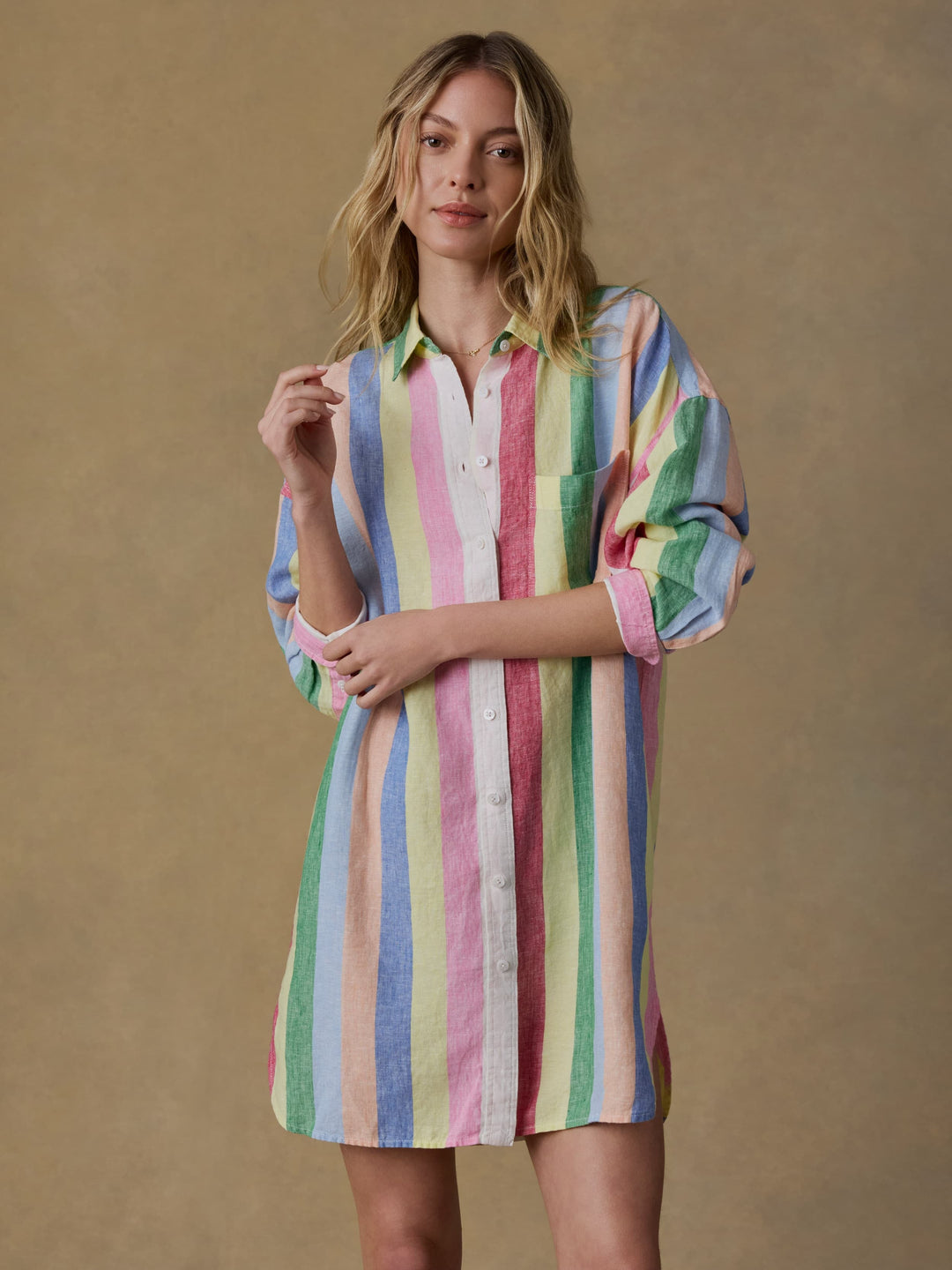 A woman wears the Zertz LAGUNA LINEN MINI SHIRTDRESS, featuring long sleeves and vertical pastel stripes in pink, yellow, green, and blue, standing against a plain light brown background.