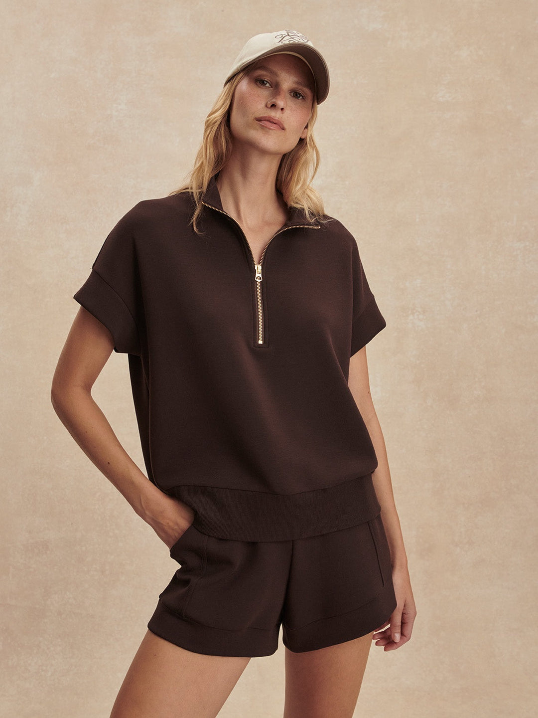 Wearing the Zertz RITCHIE SHORT SLEEVE SWEAT in coffee bean, paired with matching shorts and a tan cap, a person stands against a beige background with one hand in their pocket.