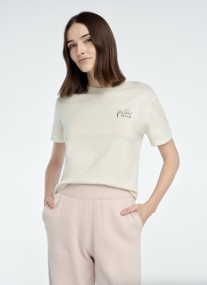 A woman with straight brown hair wears the Zertz THE FAVORITE GRAD TEE in cream and light pink sweatpants, standing with her hands in her pockets against a plain light background.