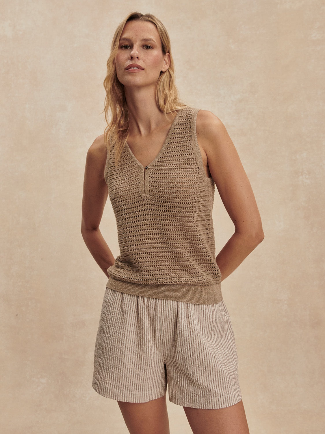 A woman stands against a neutral background in the Zertz RAMON KNIT TANK-SEPIA TWIST, a sleeveless V-neck top. Her relaxed confidence and blonde hair highlight this versatile layering piece’s fresh, effortless style with a subtle sepia hue.