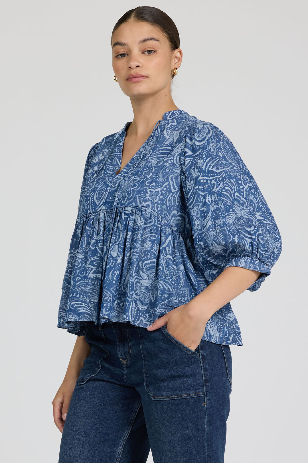 A woman wears the Zertz MALIA TOP, a loose-fitting floral blue top with three-quarter sleeves, paired with dark blue jeans, standing with one hand in her pocket against a plain light background.