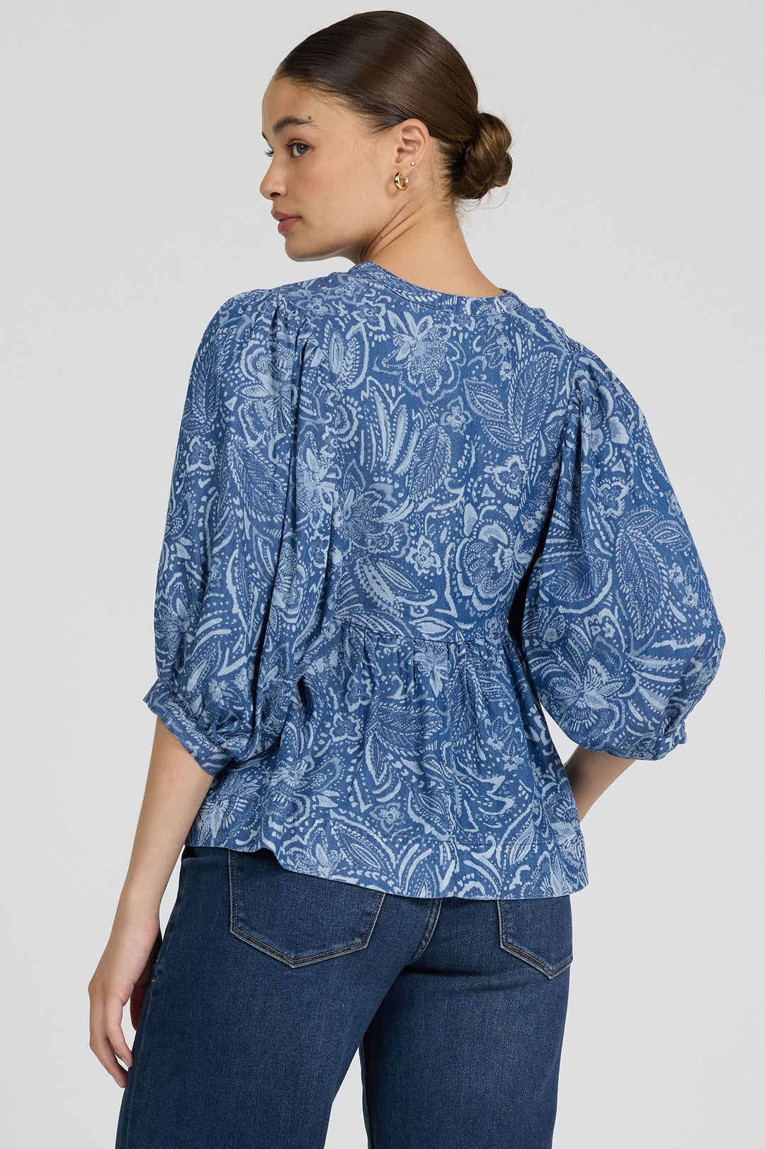 A woman with her hair in a low bun wears the Zertz MALIA TOP, a feminine blue patterned blouse with puffed sleeves made from breathable fabric, paired with dark blue jeans, as she looks over her shoulder against a plain light background.
