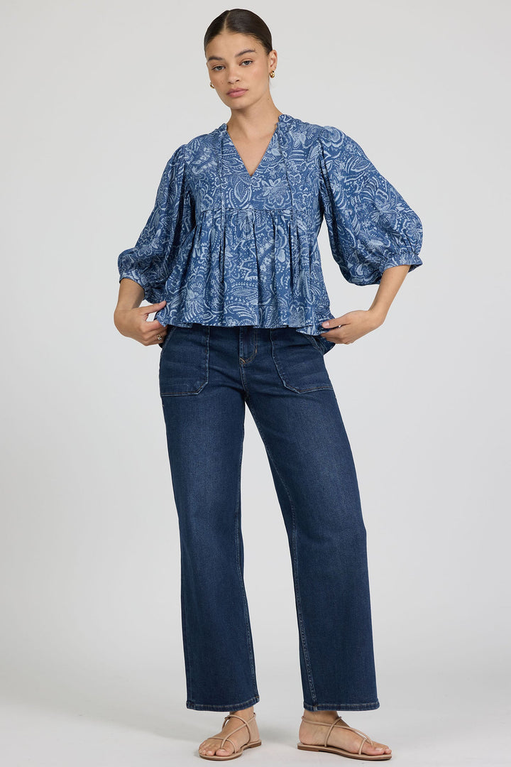 A woman wears the Zertz MALIA TOP, a feminine, loose-fitting blouse with a blue pattern and balloon sleeves, paired with dark blue straight-leg jeans and beige sandals against a plain light background.