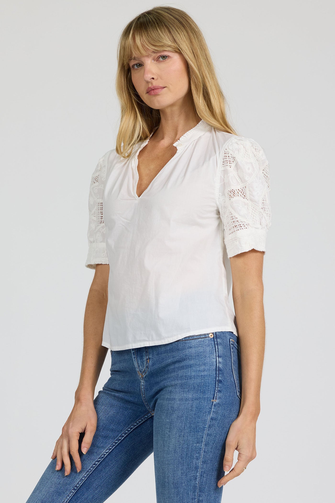 A woman with long blonde hair wears the Zertz SAMMI TOP, a versatile white blouse with lace details, paired with blue jeans against a plain light background.