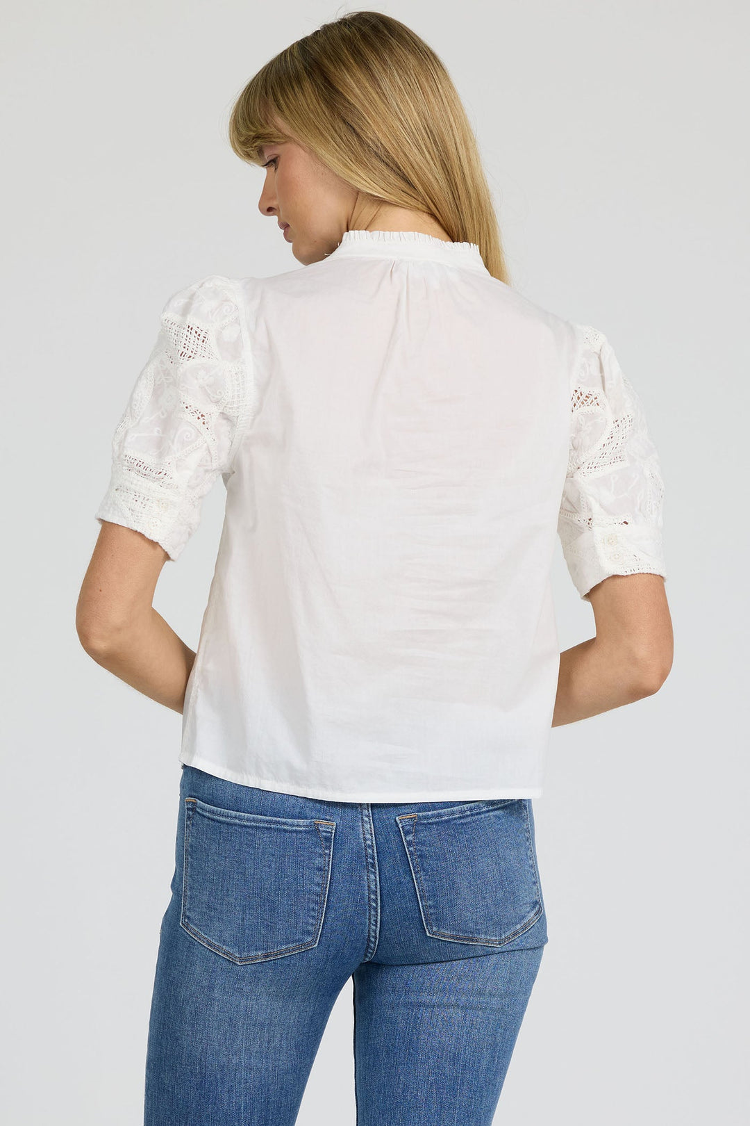 A woman with straight blonde hair is seen from the back, wearing the Zertz SAMMI TOP—a versatile white blouse with lace details—and blue jeans against a plain light background.