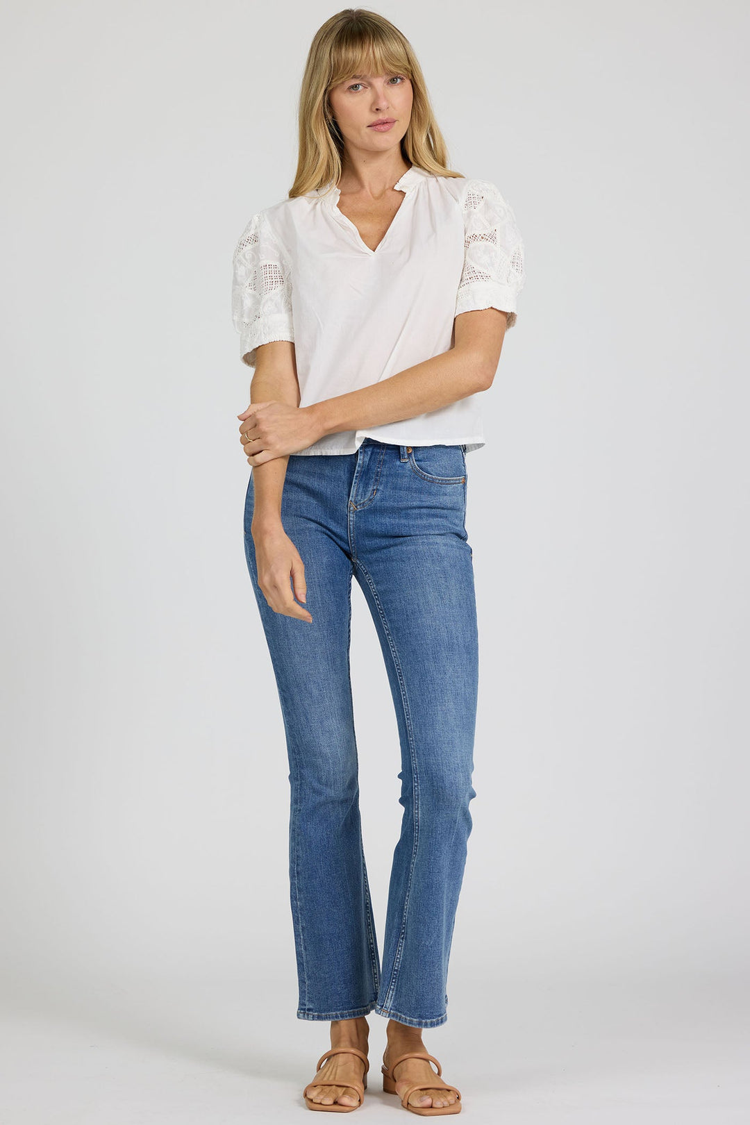 A woman with blonde hair and bangs stands against a plain background, wearing the Zertz SAMMI TOP—a white blouse with lace details—paired with blue jeans and tan sandals. She holds one arm with her hand.