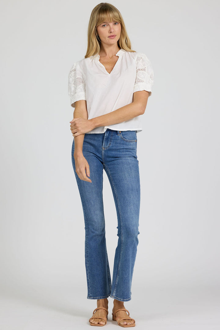A woman with blonde hair and bangs stands against a plain background, wearing the Zertz SAMMI TOP—a white blouse with lace details—paired with blue jeans and tan sandals. She holds one arm with her hand.
