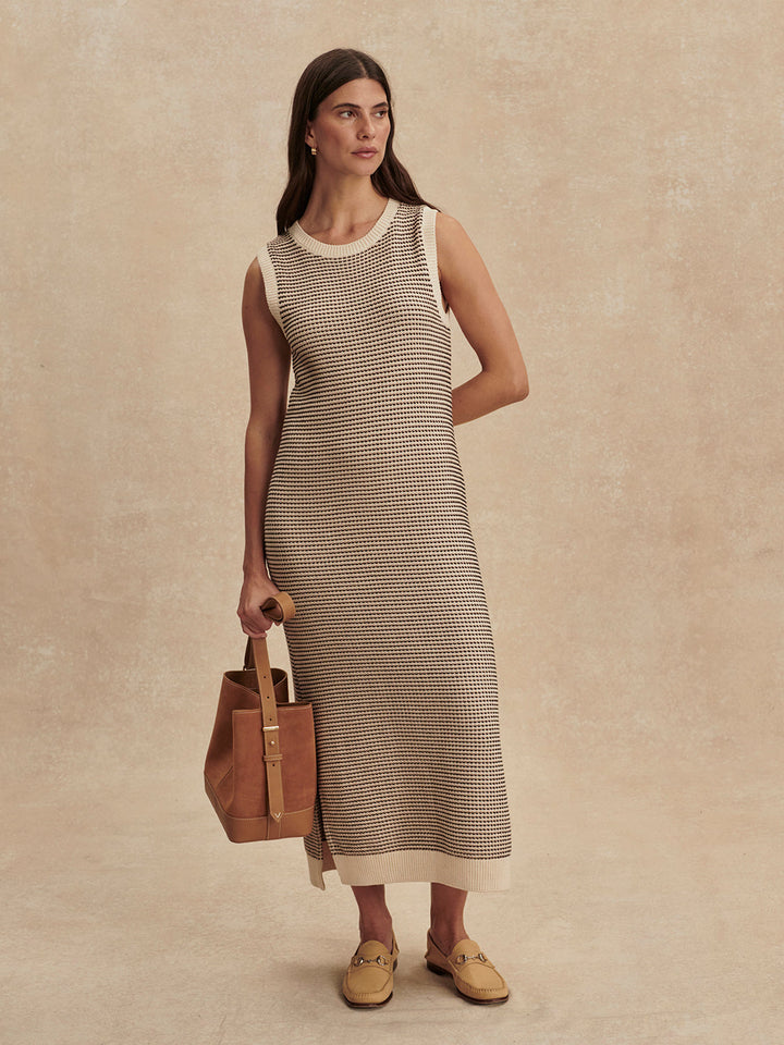 A woman with long brown hair wears the Zertz SADIA STITCH KNIT DRESS, a sleeveless piece with textured stitching, paired with tan loafers and a brown leather tote, standing against a beige background—perfect for any versatile wardrobe.