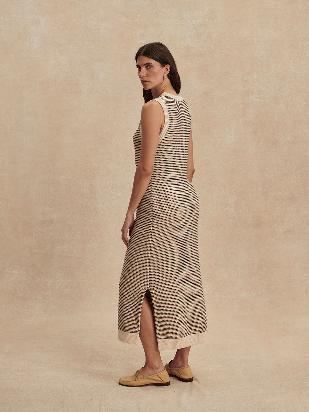 A woman with long brown hair wears the Zertz SADIA STITCH KNIT DRESS, a sleeveless, ankle-length design with beige and brown patterns and textured stitching. She stands sideways against a neutral background, tan shoes completing her look.