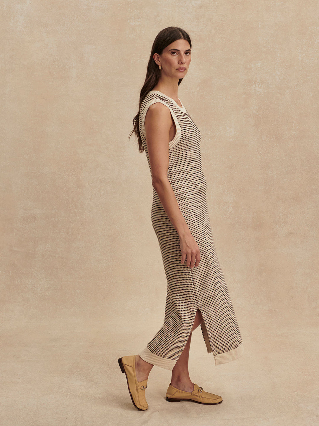 A woman with long brown hair wears the Zertz SADIA STITCH KNIT DRESS—an ankle-length, sleeveless knit dress with textured stitching—and beige loafers, standing against a neutral textured background.