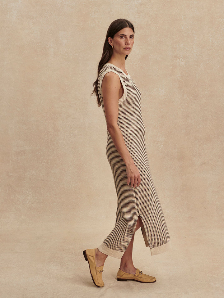 A woman with long brown hair wears the Zertz SADIA STITCH KNIT DRESS—an ankle-length, sleeveless knit dress with textured stitching—and beige loafers, standing against a neutral textured background.