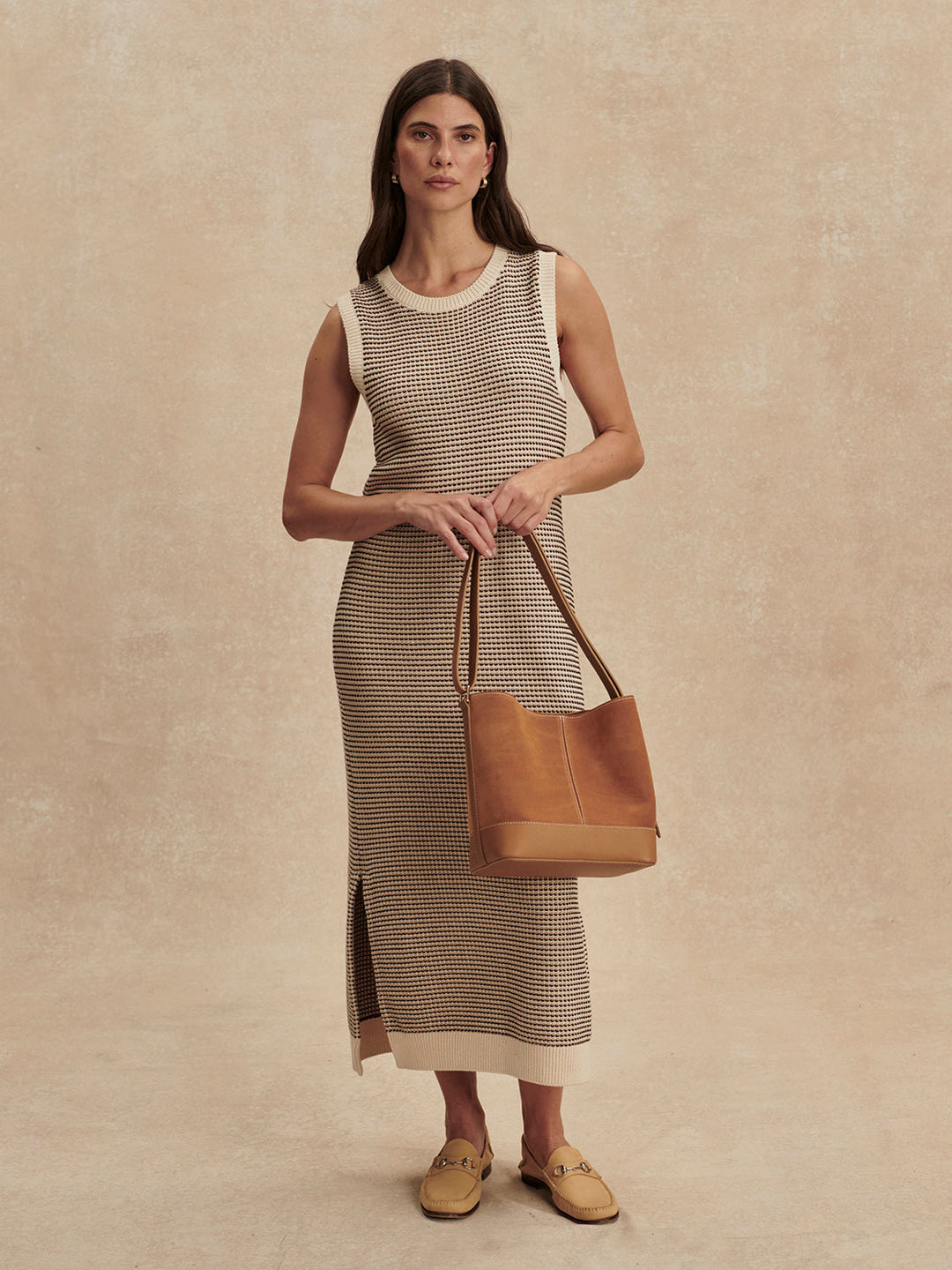 A woman stands against a neutral background wearing the Zertz SADIA STITCH KNIT DRESS—an ankle-length, sleeveless beige and brown knit dress—and holds a large light brown leather tote bag while wearing tan loafers.