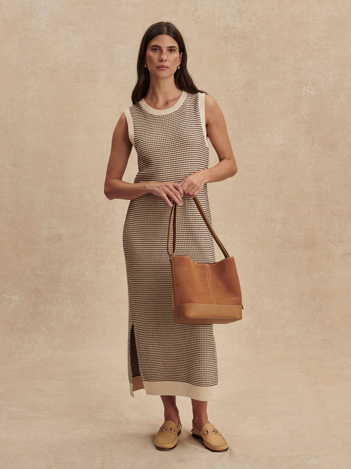 A woman stands against a neutral background wearing the Zertz SADIA STITCH KNIT DRESS—an ankle-length, sleeveless beige and brown knit dress—and holds a large light brown leather tote bag while wearing tan loafers.