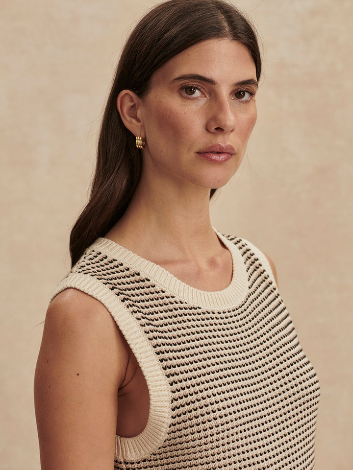 A woman with long brown hair models the Zertz SADIA STITCH KNIT DRESS—featuring black and cream stripes with textured stitching—and gold hoop earrings, standing against a neutral beige background looking to the side.