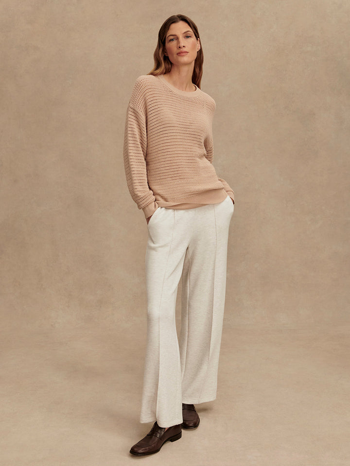 A woman wears the Zertz KERSHAW SWEATER TOP in beige with wide-legged light gray pants and brown shoes, posing against a neutral textured backdrop. This cozy knit sweater offers classic style and versatile comfort for any wardrobe.