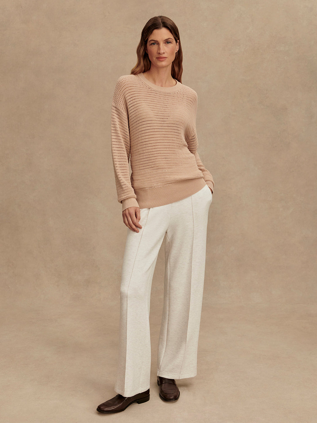 A woman stands against a beige background wearing the Zertz KERSHAW SWEATER TOP in light brown, wide-leg white trousers, and brown shoes, with her hands in her pockets.