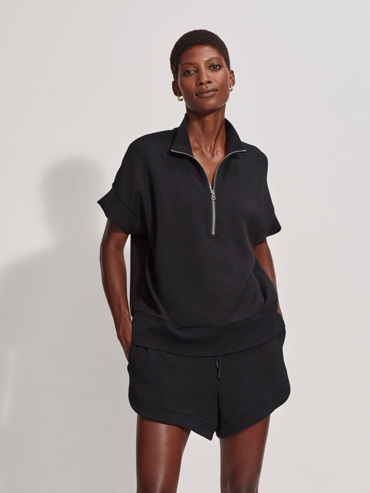 A woman with short hair and hoop earrings stands confidently against a light background in the Zertz RITCHIE SHORT SLEEV HALF ZIP TOP BLACK and matching shorts, her hands tucked into her pockets for a stylish, casual look.
