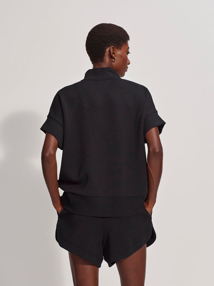 A person faces away from the camera, wearing the Zertz RITCHIE SHORT SLEEV HALF ZIP TOP BLACK with matching black shorts, hands in back pockets, set against a plain light background.