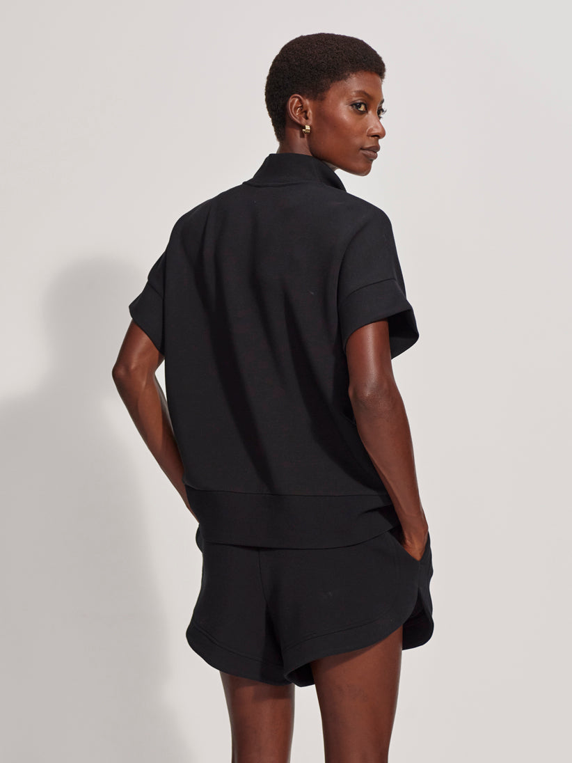 A person with short hair stands sideways, looking over their shoulder and posing with hands in pockets, wearing the Zertz RITCHIE SHORT SLEEV HALF ZIP TOP BLACK and matching shorts against a plain light background.