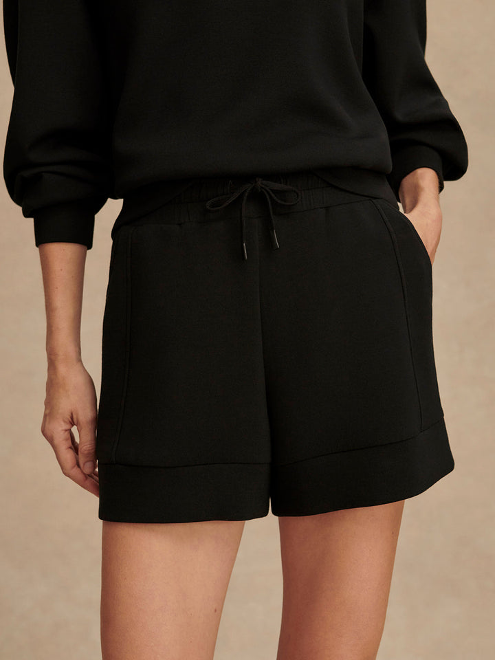 A person wears Zertz ATRIUM HIGH RISE SHORT 4.5 INSEAM BLACK and a matching top, standing with one hand in their pocket against a neutral background.